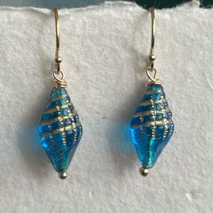 May include: A pair of dangle earrings featuring teardrop-shaped, turquoise blue glass beads with gold accents. The beads are suspended from gold-colored ear wires. The earrings have a seashell design.
