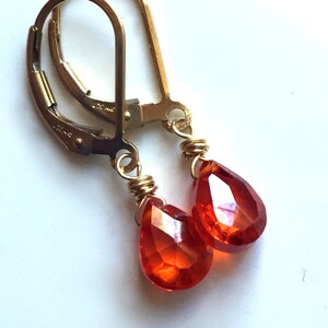 May include: A pair of gold-colored leverback earrings with teardrop-shaped, faceted orange gemstones. The gemstones are wire-wrapped in gold-colored wire. The earrings are displayed against a white background.