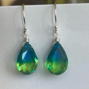 May include: A pair of dangle earrings featuring teardrop-shaped, faceted gemstones. The gems transition from a vibrant green at the base to a deep blue at the top. The earrings have silver-colored hooks and wire detailing.