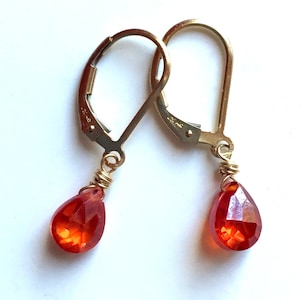 May include: A pair of gold-colored leverback earrings with teardrop-shaped, faceted, orange gemstones. The gemstones are wire-wrapped in gold-colored wire. The earrings are displayed against a white background.