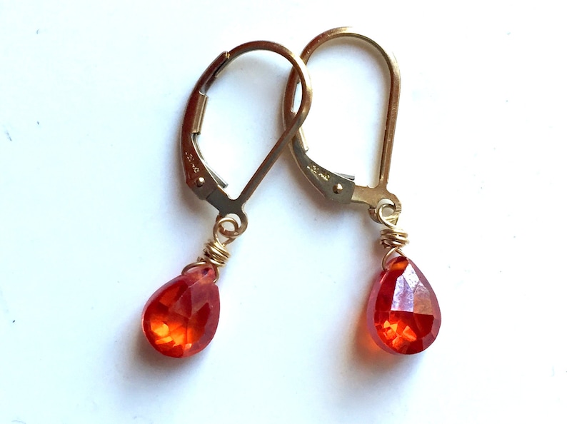 May include: A pair of gold-colored leverback earrings with teardrop-shaped, faceted, orange gemstones. The gemstones are wire-wrapped in gold-colored wire. The earrings are displayed against a white background.