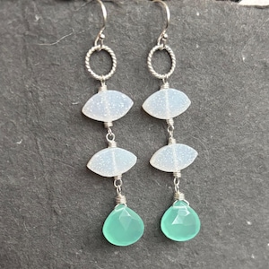 May include: Dangle earrings featuring a silver-toned twisted ring at the top, connected to two white, oval-shaped stones. Below, a teardrop-shaped, light green stone hangs. The earrings are connected with silver wire.