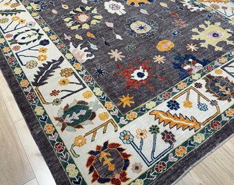 May include: A rectangular, dark gray rug with a floral pattern. The rug features a cream-colored border with colorful floral and geometric designs. The central field of the rug is filled with various floral motifs in shades of yellow, blue, and red.
