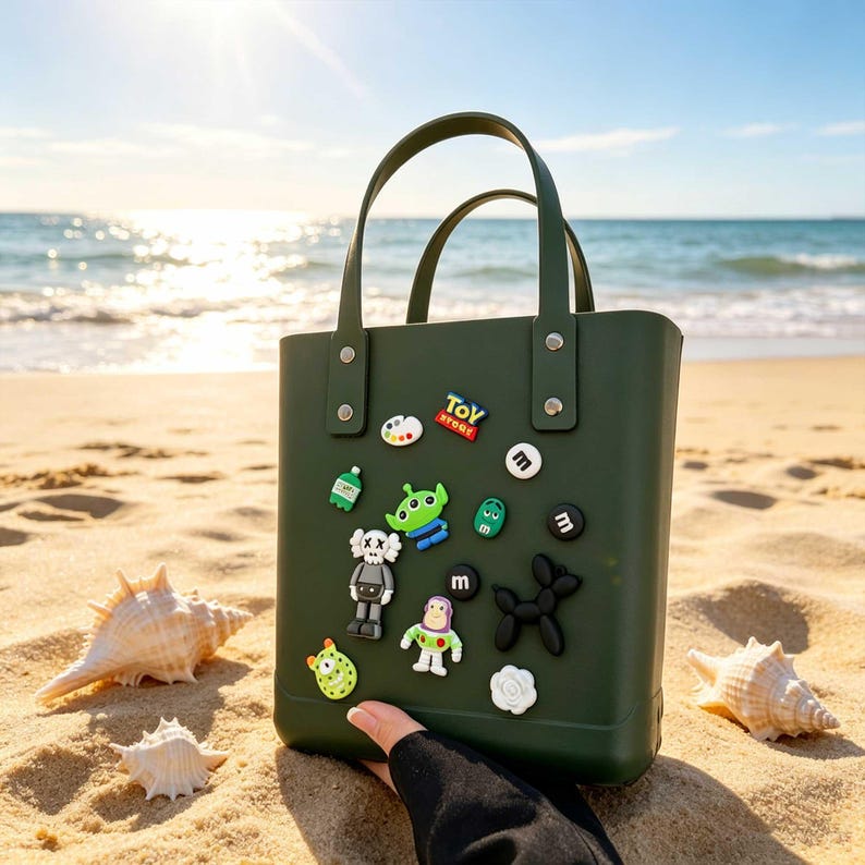 May include: A dark green tote bag with attached decorative pins, including a Toy Story logo, alien, and other cartoon characters. The bag has two handles and is on a sandy beach with seashells.