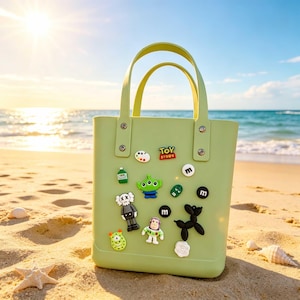 May include: A light green tote bag with two handles, decorated with colorful cartoon character charms. The bag is on a sandy beach with the ocean and sky in the background. The charms include a green alien, a robot, and the words "Toy Story".