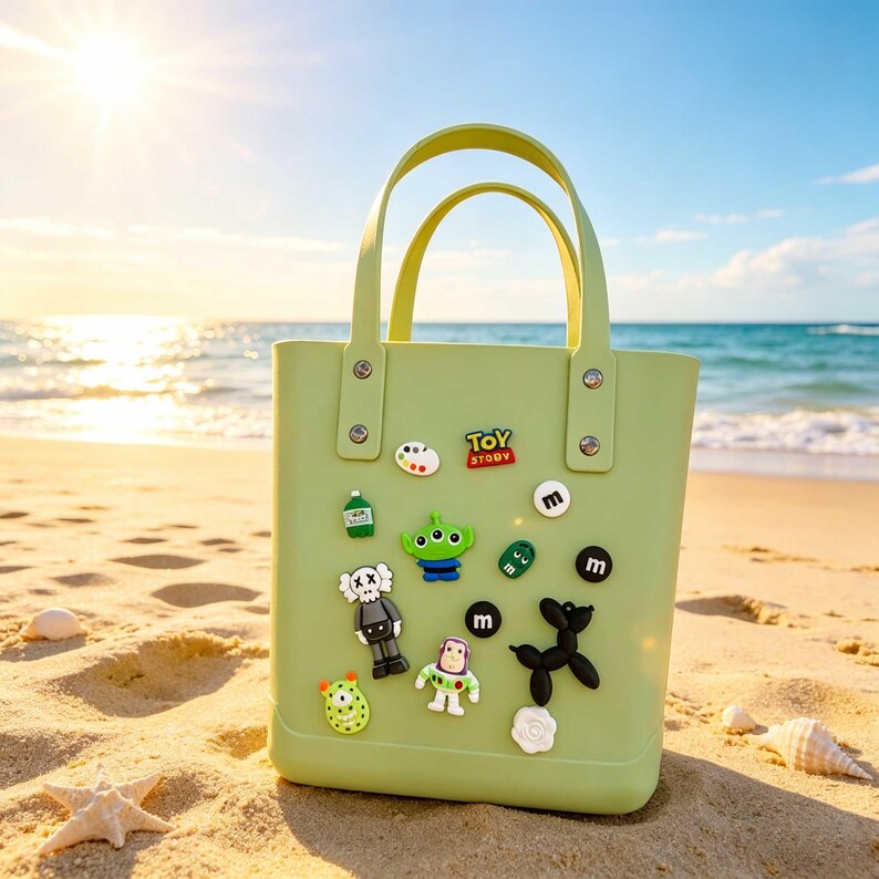 May include: A light green tote bag with two handles, decorated with colorful cartoon character charms. The bag is on a sandy beach with the ocean and sky in the background. The charms include a green alien, a robot, and the words "Toy Story".