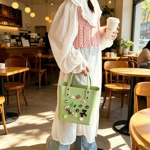May include: A light green tote bag with a rectangular shape and two handles. The bag is decorated with various colorful charms. The person is wearing a white dress and a pink sweater.