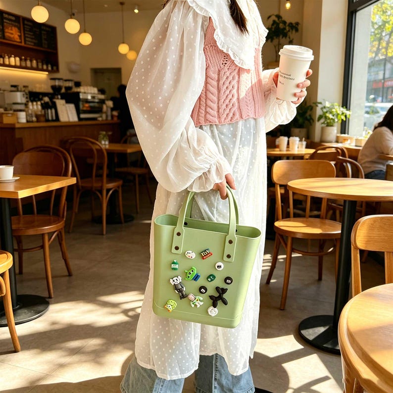 May include: A light green tote bag with a rectangular shape and two handles. The bag is decorated with various colorful charms. The person is wearing a white dress and a pink sweater.