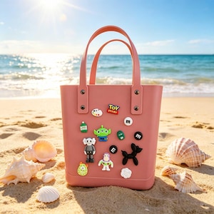 May include: A pink tote bag with two handles, decorated with colorful cartoon character charms. The bag is on a sandy beach with seashells, the ocean, and a bright sun in the background.