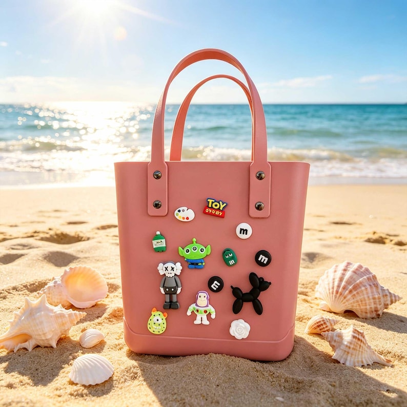 May include: A pink tote bag with two handles, decorated with colorful cartoon character charms. The bag is on a sandy beach with seashells, the ocean, and a bright sun in the background.