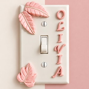 Op de afbeelding: Een rechthoekige gebroken witte lichtschakelaarplaat met twee roze gebeeldhouwde bladeren aan de linkerkant en de naam OLIVIA in verhoogde roze hoofdletters aan de rechterkant, voorzien van een centrale witte tuimelschakelaar en twee zilveren schroeven.