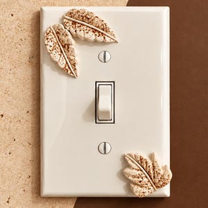 May include: A rectangular off-white light switch wall plate featuring three raised, textured leaf-shaped embellishments in a speckled tan and brown finish. The plate includes a central toggle switch and two silver-toned mounting screws.