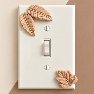 May include: A rectangular white light switch cover plate featuring three attached decorative leaf-shaped elements with a speckled tan texture. The plate includes a central toggle switch and two silver-colored mounting screws.