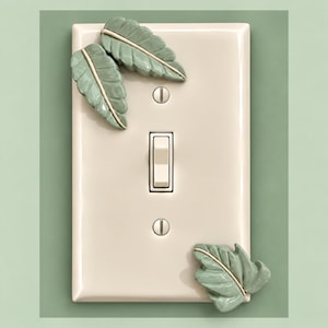 May include: A rectangular cream-colored light switch plate featuring three dimensional sage green ceramic leaves attached to the surface. The plate includes a central toggle switch and two silver screws for mounting, with two leaves at the top left and one leaf at the bottom right.