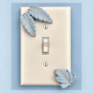 Op de afbeelding: Een rechthoekige, crèmekleurige lichtschakelaar-muurplaat met drie gebeeldhouwde, lichtblauwe bladaccenten. De plaat bevat een centrale tuimelschakelaar en twee zilverkleurige schroeven boven en onder het schakelmechanisme.