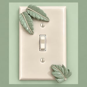 Op de afbeelding: Een rechthoekige, gebroken witte lichtschakelaar-muurplaat met drie opgeplakte, gevormde saliegroene keramische bladeren. De plaat is voorzien van een centrale tuimelschakelaar en twee platte schroeven voor montage, met een glanzende afwerking.