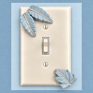 Op de afbeelding: Een rechthoekige, crèmekleurige wandplaat voor een lichtschakelaar met drie gevormde, lichtblauwe keramische bladeren op het oppervlak. De plaat bevat een centrale tuimelschakelaar en twee bijpassende schroeven voor installatie.