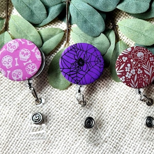 May include: Five Halloween-themed retractable badge holders. Designs include skulls, spiderwebs, floral patterns, jack-o'-lanterns, and a skull with a coffin. The badge reels are round with a clear plastic clip and a metal belt clip.