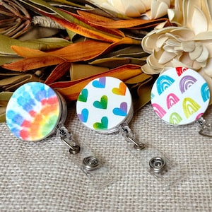 May include: Three retractable badge holders with colorful designs. One has a tie-dye pattern, another features hearts in various colors, and the third displays rainbow arches. Each holder has a clear plastic strap and a metal clip.