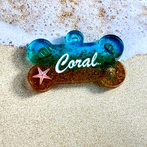 May include: A blue and gold dog bone shaped tag with the word "Coral" written in white. The tag has a starfish, a seashell, and a small fish embedded in the resin.