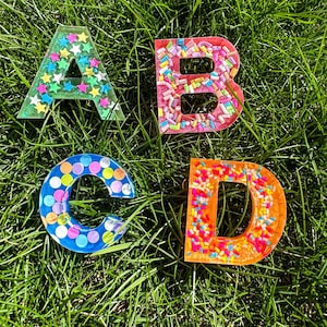 May include: Four resin letters, A, B, C, and D, with different colourful embellishments. The letters are on a green grassy surface.