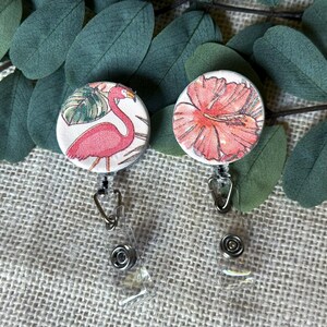 May include: Two round retractable badge holders. One features a pink flamingo with tropical leaves, the other a pink hibiscus flower. Both have clear plastic clips and silver-colored metal hardware. The background includes green leaves and a textured beige surface.