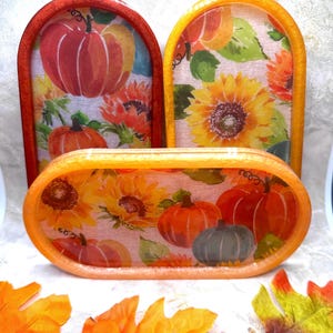 May include: Three oval-shaped resin trays with a fall theme. The trays are decorated with a white background and a pattern of pumpkins and sunflowers. The trays are in shades of orange, yellow, and red.