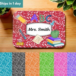 Op de afbeelding: Een rode bureaumat met compositieboekthema met de tekst "Mrs. Smith" in een wit label. De mat bevat schoolgerelateerde illustraties, waaronder een potlood, appel, boeken en de letters "ABC".