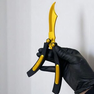 May include: A butterfly knife with a gold blade and accents, and black handles. The knife is held against a white background. The blade has a curved shape.