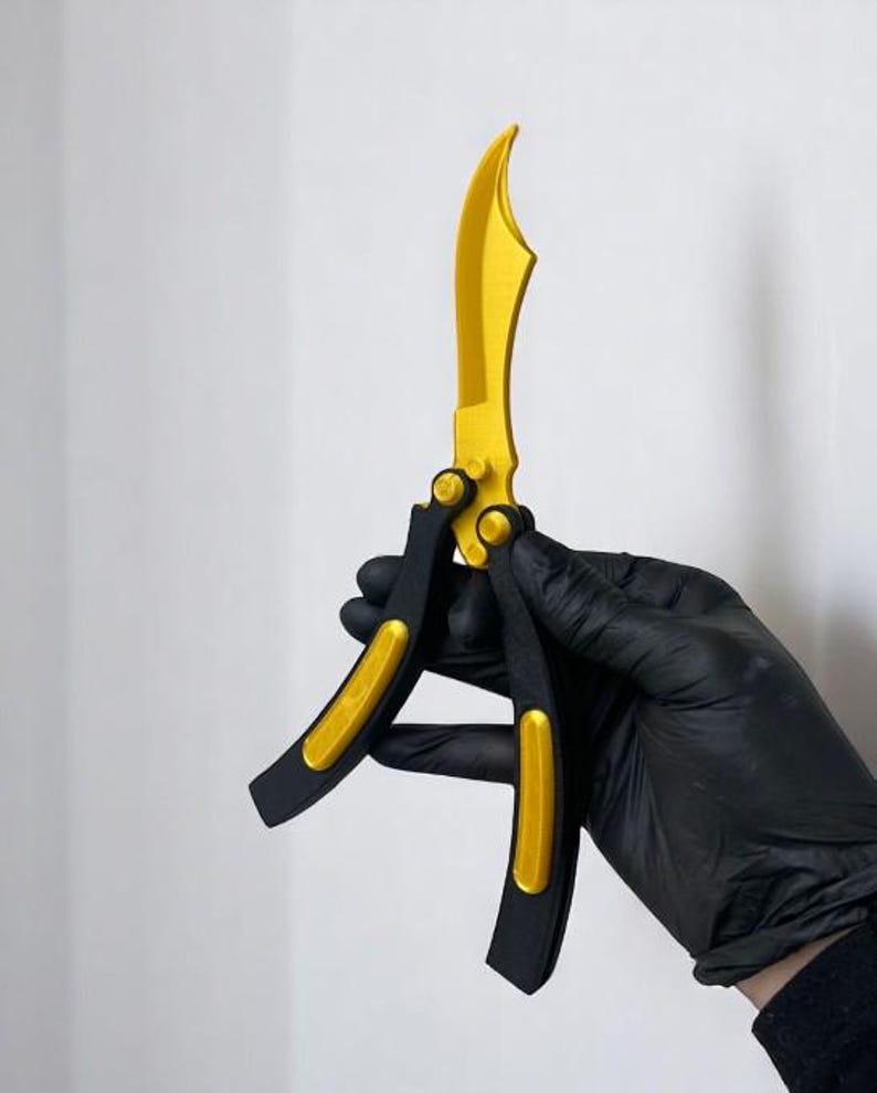 May include: A butterfly knife with a gold blade and accents, and black handles. The knife is held against a white background. The blade has a curved shape.