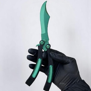 May include: A green and black butterfly knife with a curved blade. The handle is black with green accents. The knife is held against a white background.