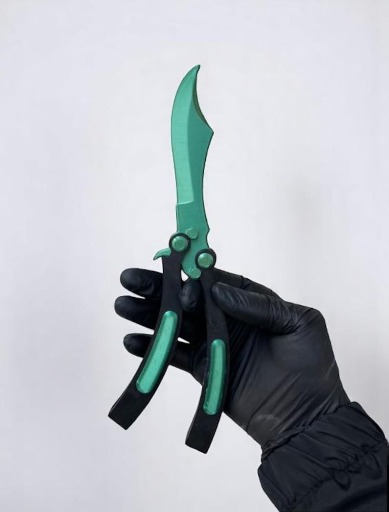 May include: A green and black butterfly knife with a curved blade. The handle is black with green accents. The knife is held against a white background.