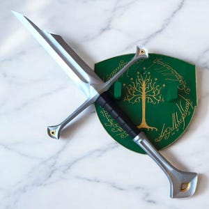 May include: A silver sword with a black handle and a green shield with a gold tree and inscription. The inscription reads "One Ring to rule them all, One Ring to find them, One Ring to bring them all and in the darkness bind them."