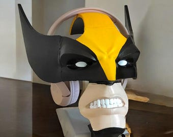 May include: A 3D printed headphone stand shaped like Wolverine's head. The stand is black and yellow with white eyes and a snarling mouth. The headphones are white and are resting on the stand's ears.