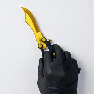 May include: A gold-coloured butterfly knife with a curved blade and black handle. The knife is held by a gloved hand against a white background. The blade has a curved shape.