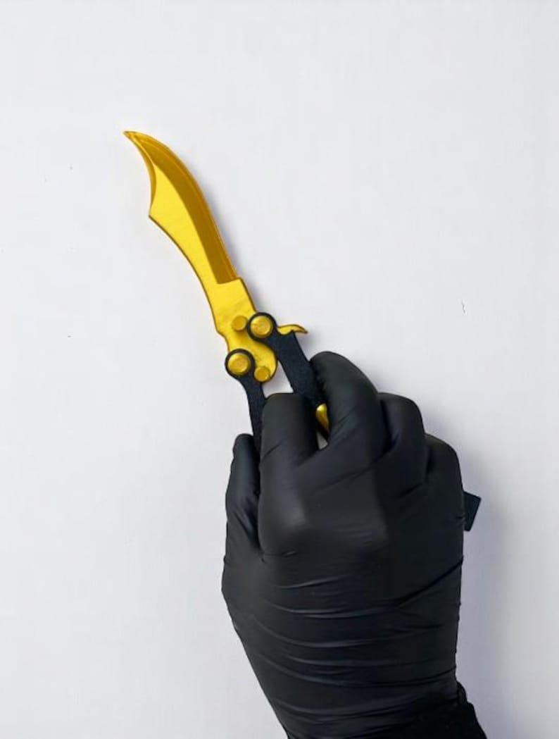 May include: A gold-coloured butterfly knife with a curved blade and black handle. The knife is held by a gloved hand against a white background. The blade has a curved shape.