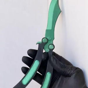 May include: A green and black butterfly knife, set against a white backdrop. The blade and accents are a vivid green, contrasting with the black handle. The knife is a folding style, with a curved blade.
