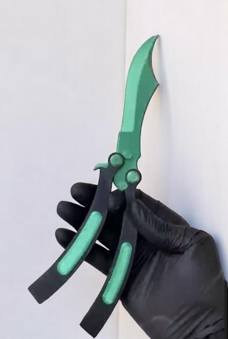 May include: A green and black butterfly knife, set against a white backdrop. The blade and accents are a vivid green, contrasting with the black handle. The knife is a folding style, with a curved blade.