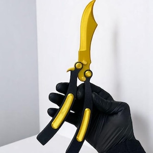 May include: A gold and black butterfly knife. The blade is gold, and the handle is black with gold accents. The knife is held in a black gloved hand against a white background.