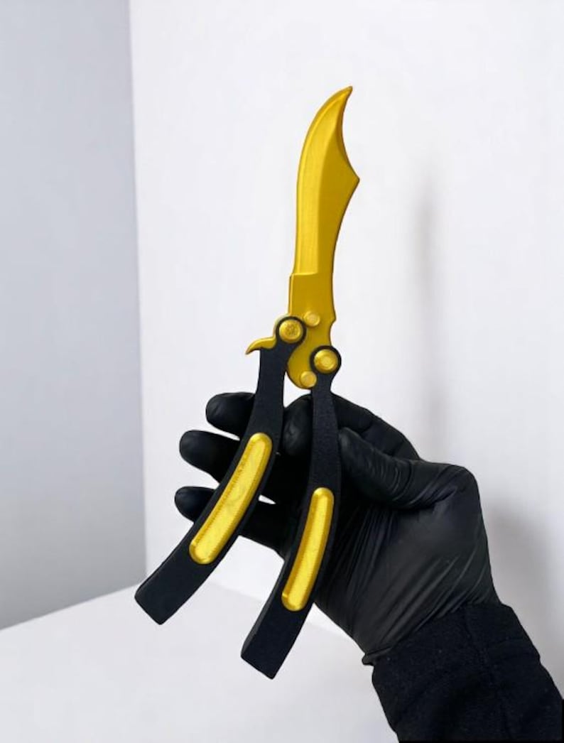 May include: A gold and black butterfly knife. The blade is gold, and the handle is black with gold accents. The knife is held in a black gloved hand against a white background.
