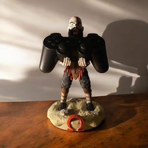 Könnte beinhalten: Figur eines kahlen, bärtigen Kriegers, der einen großen schwarzen Videospiel-Controller hält. Die Figur steht auf einem beigen Sockel mit einem roten Omega-Symbol. Der Krieger trägt braune und schwarze Kleidung.
