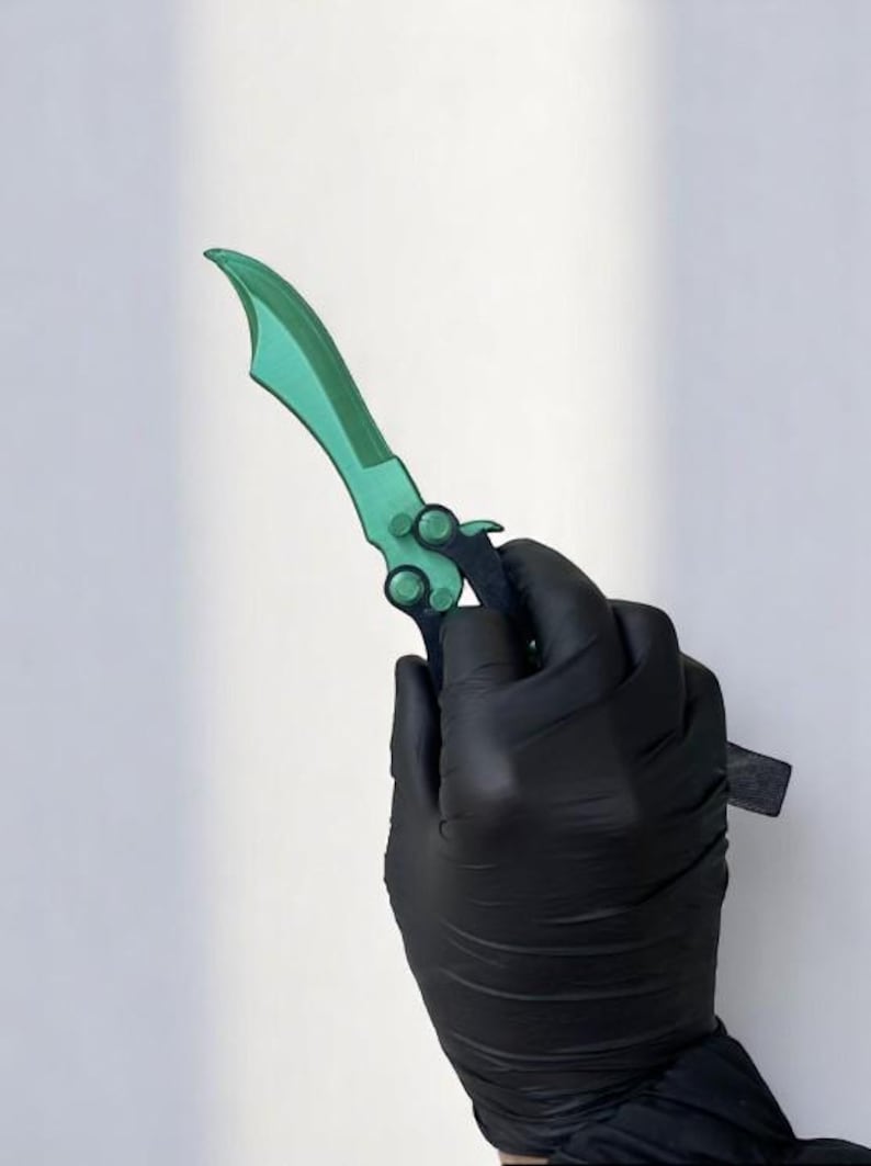 May include: A green and black butterfly knife with a curved blade. The blade is a vibrant green, and the handle is black. The knife is held by a gloved hand against a white background.