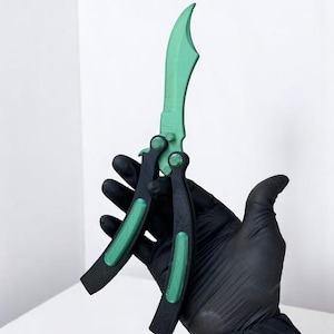 May include: A green and black butterfly knife. The blade is a vibrant green, and the handle is black with green accents. The knife is held by a gloved hand against a white background. The knife appears to be made of plastic.