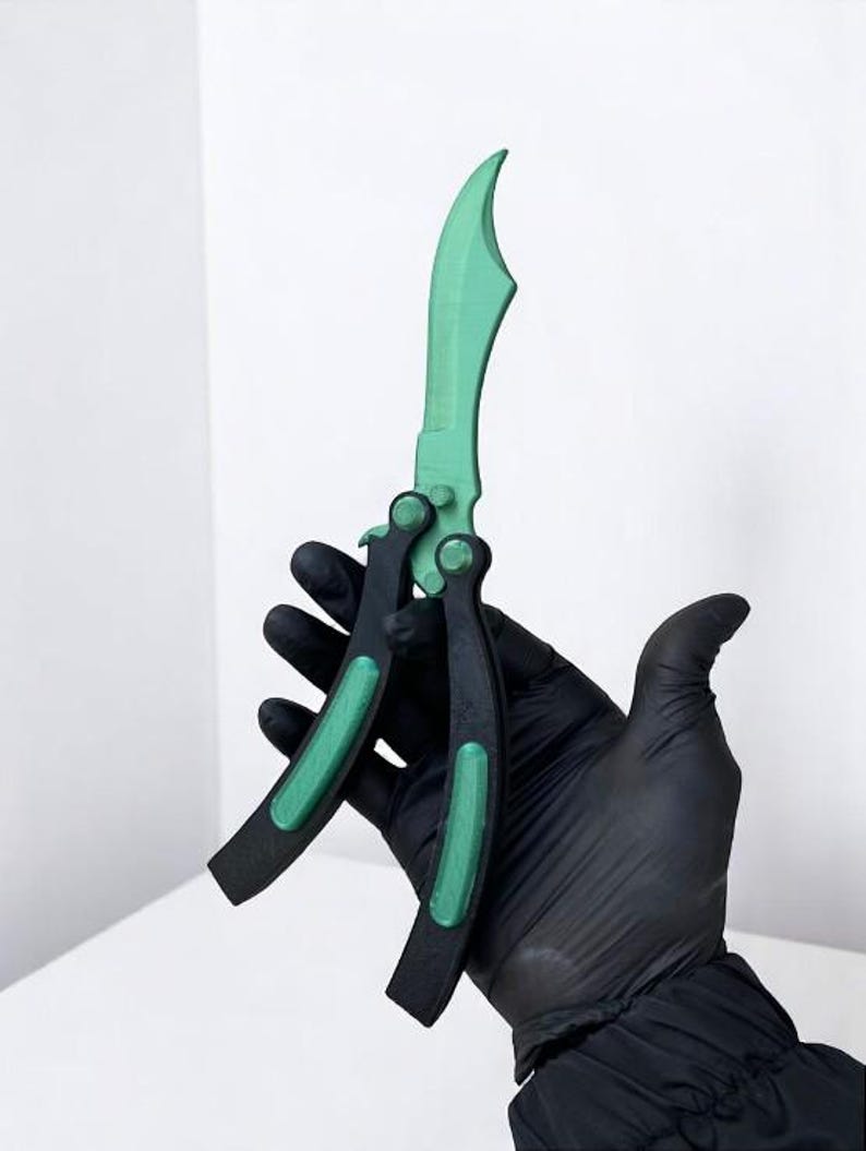 May include: A green and black butterfly knife. The blade is a vibrant green, and the handle is black with green accents. The knife is held by a gloved hand against a white background. The knife appears to be made of plastic.
