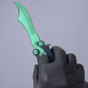 May include: A green butterfly knife with a curved blade and a black handle. The knife is held in a gloved hand against a grey background. The blade has a unique shape.