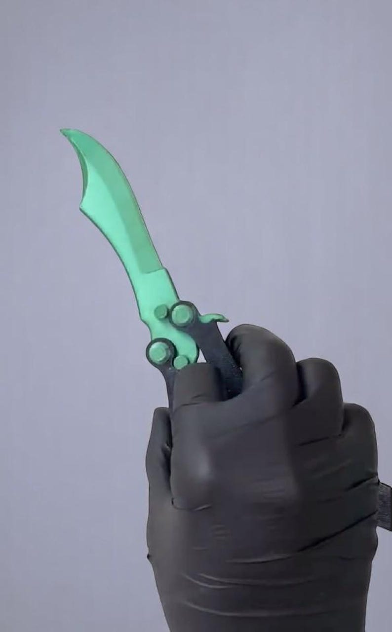 May include: A green butterfly knife with a curved blade and a black handle. The knife is held in a gloved hand against a grey background. The blade has a unique shape.