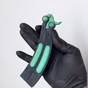 May include: A butterfly knife with a black handle and green accents. The handle is split into two parts, with a green section on each side. The top of the knife has a green design. The knife is held by a gloved hand.