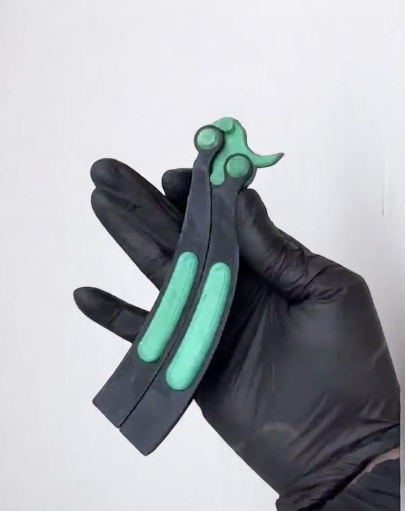 May include: A butterfly knife with a black handle and green accents. The handle is split into two parts, with a green section on each side. The top of the knife has a green design. The knife is held by a gloved hand.