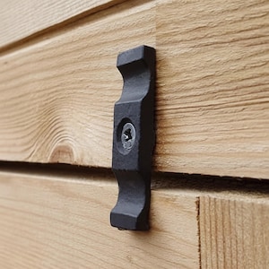 Puede incluir: Un sujetador de metal negro mate con una forma vertical contorneada y un orificio central para tornillo, fijado a una superficie de madera marrón claro con un tornillo plateado. El sujetador presenta un acabado texturizado y diseño simétrico.