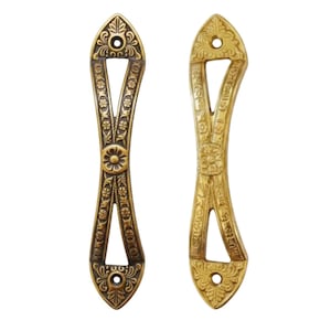 May include: Two elongated, vertical metal door handles with a pointed top and bottom. The left handle features a dark, antique brass finish with intricate floral and leaf engravings. The right handle has a bright, polished gold-tone finish.